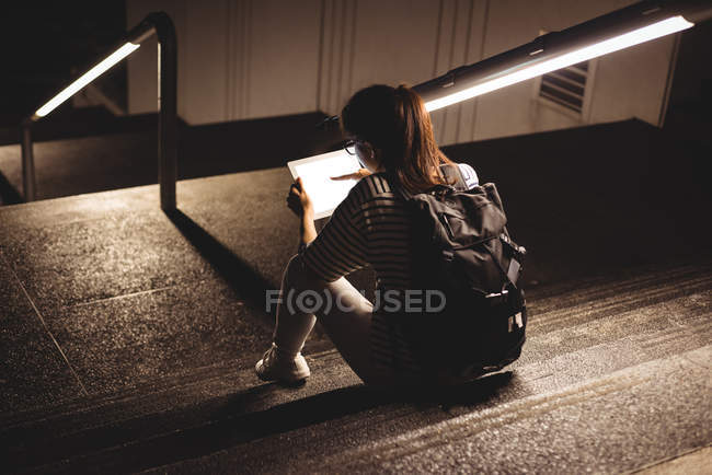 Rückansicht einer jungen Frau, die nachts im Treppenhaus sitzt und ein digitales Tablet benutzt — Stockfoto