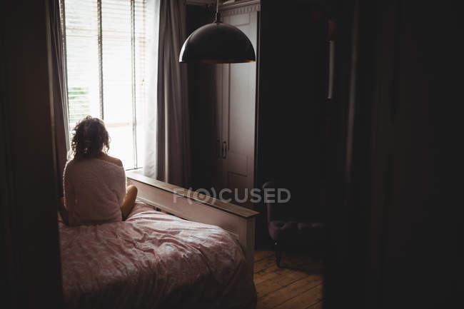 Rückansicht einer Frau, die morgens auf dem Bett sitzt und durch das Fenster schaut — Stockfoto