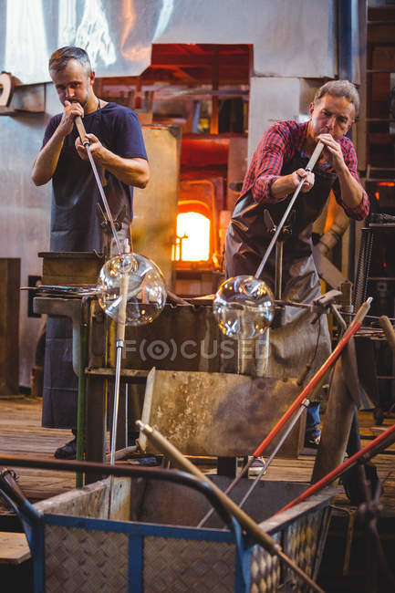 Sopladores de vidrio dando forma a un vidrio en las tuberías de soplado en la fábrica de soplado de vidrio - foto de stock