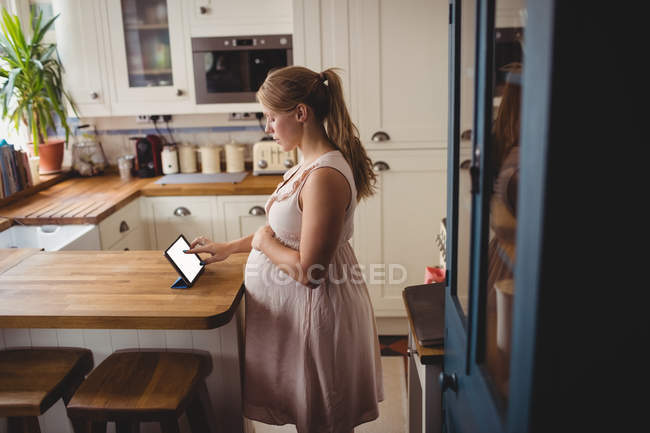 Vue latérale de la femme enceinte utilisant une tablette numérique dans la cuisine à la maison — Photo de stock