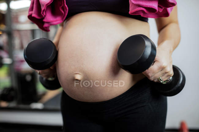 Sección media de la mujer embarazada levantando pesas en el gimnasio - foto de stock