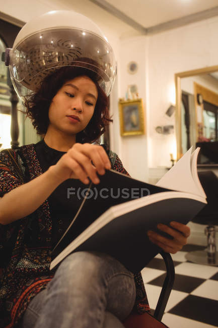 Stylish woman reading a magazine while sitting under a hairdryer at hair salon — Stock Photo