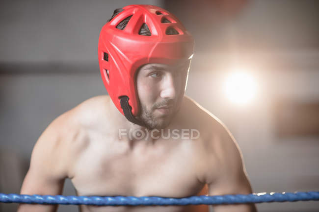 Portrait of Male boxer in protective boxing helmet leaning on ropes of boxing ring at fitness studio — Stock Photo