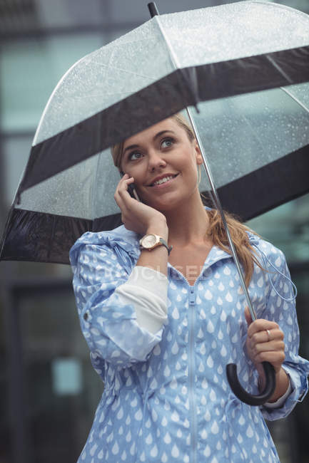 Schöne Frau mit Regenschirm, während sie auf der Straße mit dem Smartphone spricht — Stockfoto