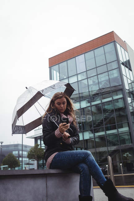 Красивая женщина, держащая зонтик и используя смартфон на улице — стоковое фото