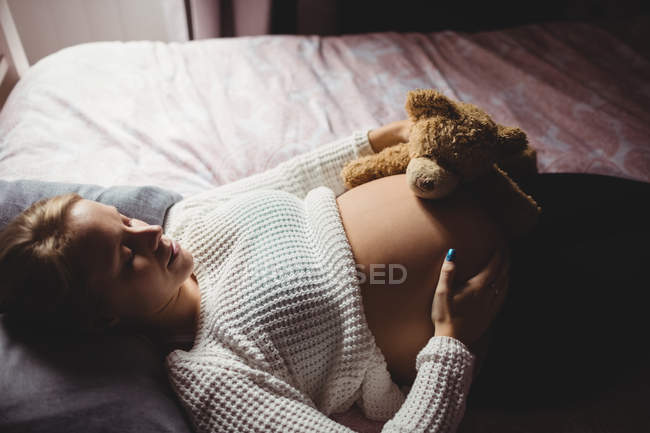 Vista de ángulo alto de la mujer embarazada sosteniendo oso de peluche en el estómago mientras duerme en el dormitorio en casa - foto de stock