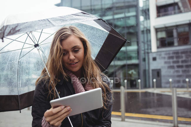 Красива жінка тримає парасольку і використовує цифровий планшет на вулиці — стокове фото