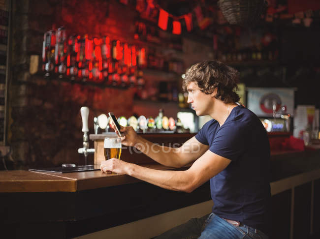 Hombre con vaso de cerveza usando teléfono móvil en el mostrador en el bar - foto de stock