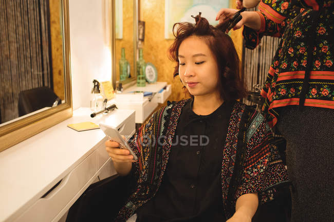 Женщина, использующая мобильный телефон, выпрямляет волосы в парикмахерской — стоковое фото