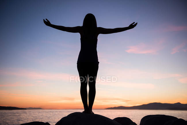 Rückansicht einer Frau, die in der Abenddämmerung Yoga auf einem Felsen macht — Stockfoto