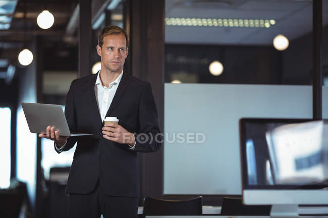 Businessman standing with a laptop and coffee cup in office — Stock Photo