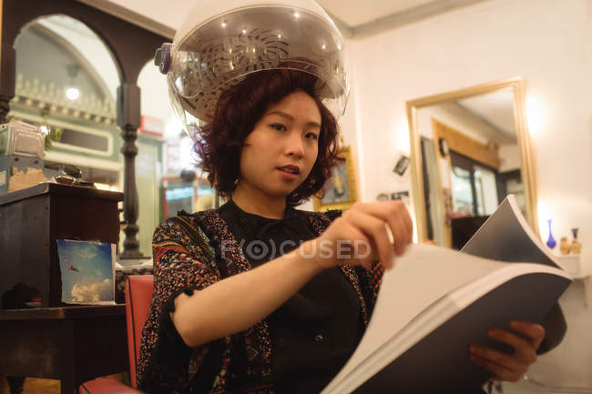 Stylish woman reading a magazine while sitting under a hairdryer at hair salon — Stock Photo
