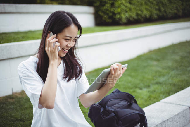 Молода жінка розмовляє на мобільному телефоні, використовуючи цифровий планшет в саду — стокове фото