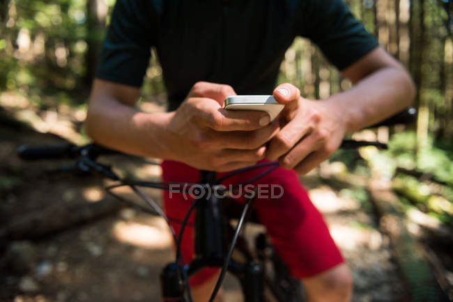 Sección media del ciclista masculino usando teléfono móvil en el bosque a la luz del sol - foto de stock