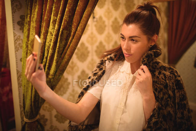Mulher tirando selfie do celular enquanto tenta uma jaqueta de pele na loja boutique — Fotografia de Stock