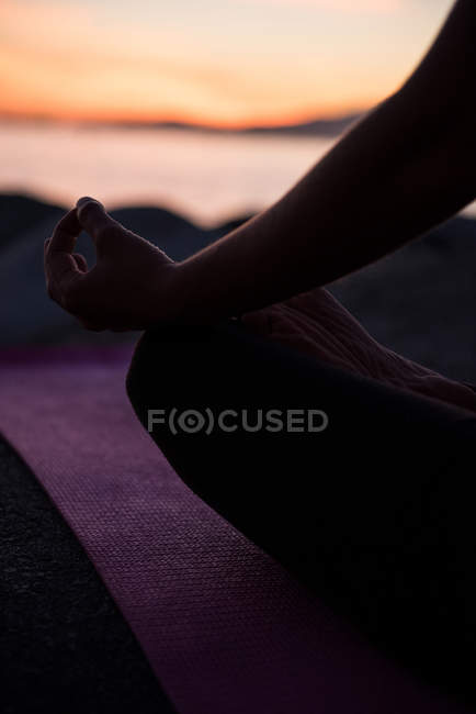 Imagem cortada de mulher sentada em posição de lótus com gesto mudra na praia ao entardecer — Fotografia de Stock