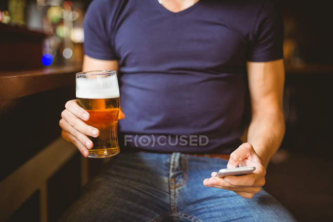 Sezione media dell'uomo che utilizza il telefono cellulare mentre ha un bicchiere di birra al bar — Foto stock