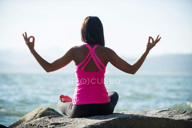 Вид ззаду жінки, що практикує йогу на скелі в сонячний день і показує жест мудра — стокове фото