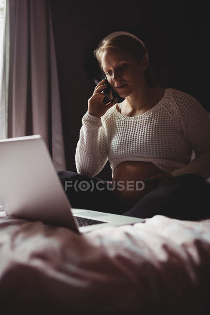 Foco seletivo da mulher grávida falando por smartphone enquanto usa laptop no quarto em casa — Fotografia de Stock