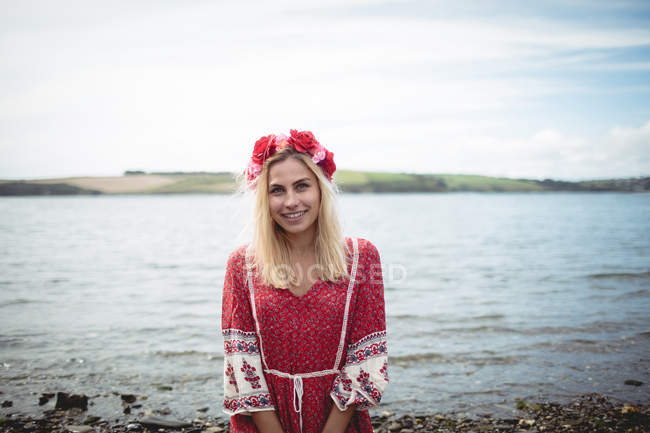 Carefree blonde woman in red dress and flower tiara standing near river and looking at camera — Stock Photo