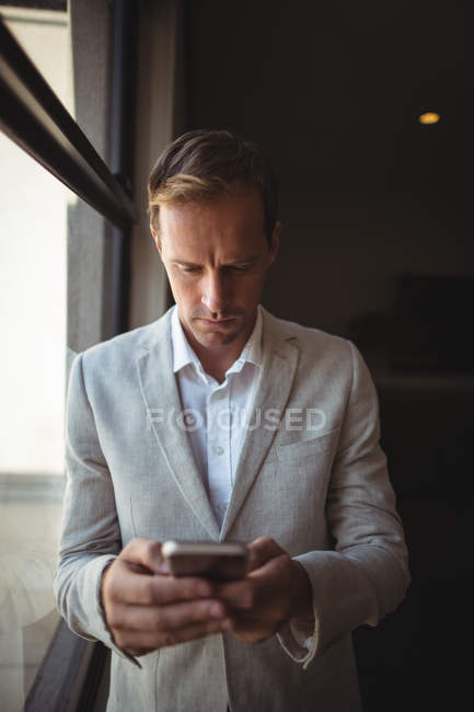Homme d'affaires pensif utilisant son téléphone portable près de la fenêtre dans le bureau — Photo de stock