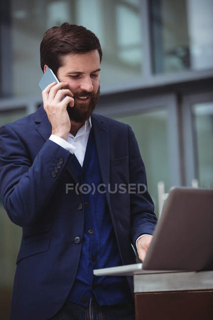 Бизнесмен разговаривает по мобильному телефону во время использования ноутбука — стоковое фото