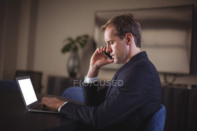 Businessman talking on the phone while using laptop in office — Stock Photo