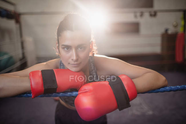 Portrait de boxeuse en gants de boxe penchée sur corde de boxe à la salle de fitness, rétroéclairée sur fond — Photo de stock