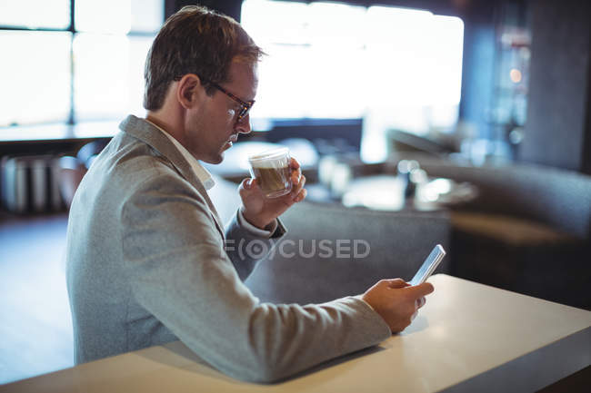 Empresário usando telefone celular enquanto toma café no café — Fotografia de Stock