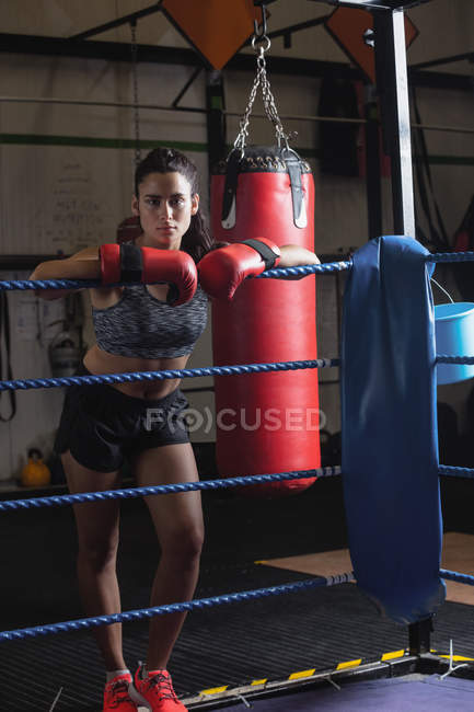 Портрет женщины-боксера в боксёрских перчатках, опирающейся на канат для бокса в фитнес-студии — стоковое фото