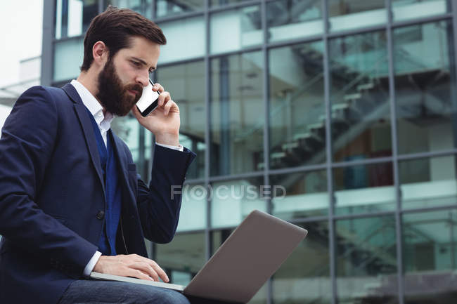 Geschäftsmann telefoniert mit Handy, während er Laptop draußen benutzt — Stockfoto