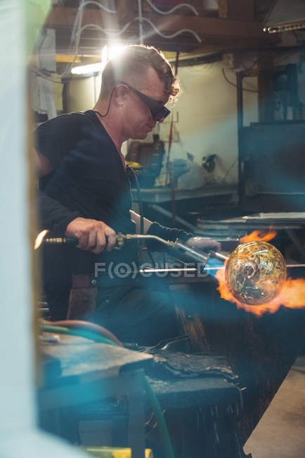 Soffiatore di vetro dando tocco finale al pezzo di vetro con la torcia soffiante in fabbrica di soffiaggio del vetro — Foto stock