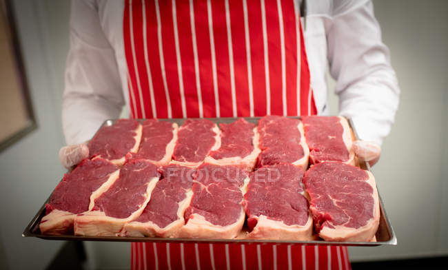 Partie médiane du boucher tenant un plateau de steaks à la boucherie — Photo de stock