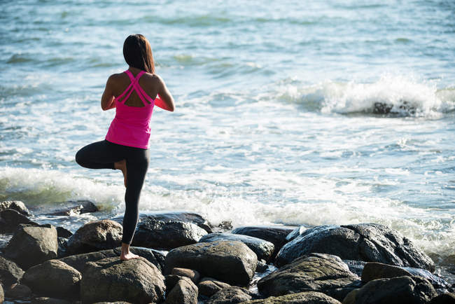 Вид ззаду жінки, що виконує йогу на скелі в сонячний день — стокове фото