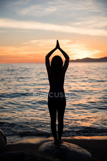 Задний вид женщины, практикующей йогу на скале на пляже во время заката — стоковое фото