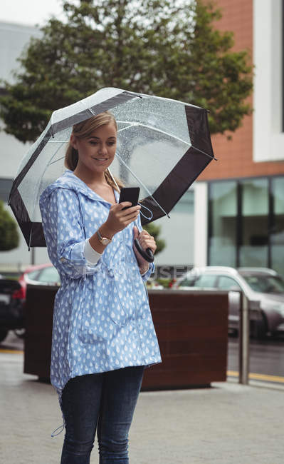Attractive woman holding umbrella while using smartphone during rainy season — Stock Photo