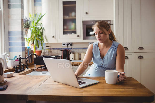 Вагітна жінка використовує ноутбук, маючи каву на кухні вдома — стокове фото