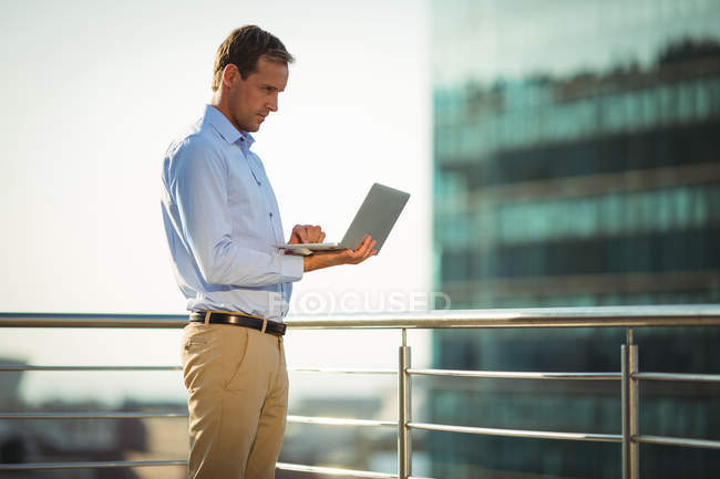 Homme d'affaires utilisant un ordinateur portable tout en se tenant sur le balcon au bureau — Photo de stock