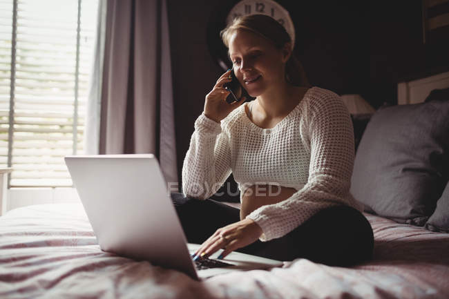 Вагітна жінка розмовляє по смартфону, використовуючи ноутбук у спальні вдома — стокове фото