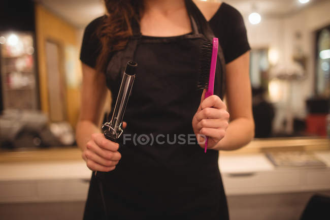 Mid-section of female hairdresser holding hair curler machine and hairbrush at saloon — Stock Photo