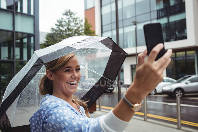 Hermosa mujer sosteniendo paraguas mientras toma selfie durante la temporada de lluvias - foto de stock
