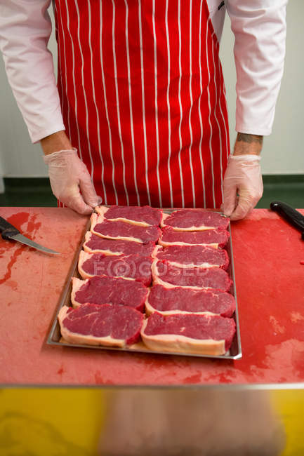Partie médiane du boucher debout au comptoir avec un plateau de steaks à la boucherie — Photo de stock