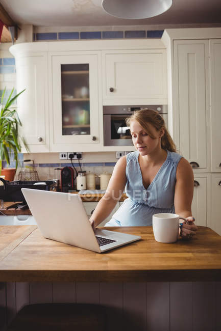 Вагітна жінка використовує ноутбук, маючи каву на кухні вдома — стокове фото