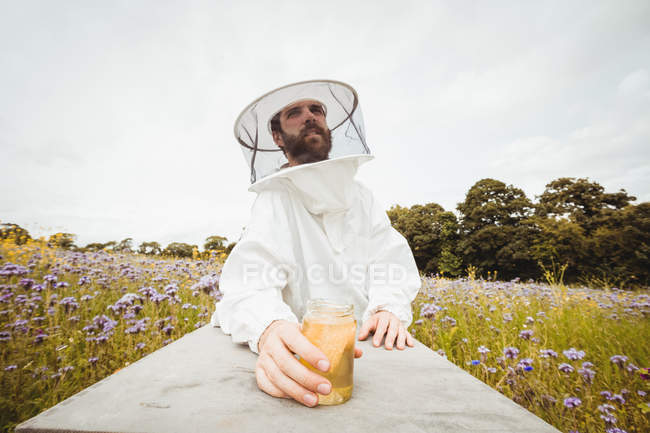 Apiculteur tenant une bouteille de miel sur une ruche dans un champ — Photo de stock