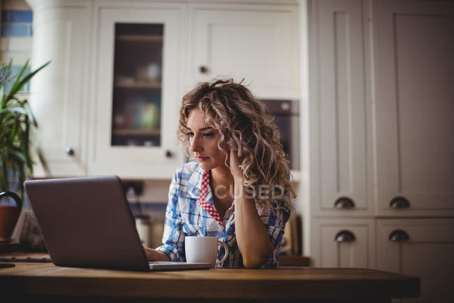 Beautiful woman using laptop while having coffee in kitchen at home — Stock Photo