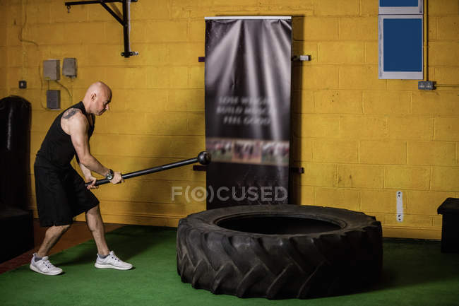 Vue latérale du boxeur thaïlandais frappant le pneu avec le marteau de traîneau dans la salle de fitness — Photo de stock