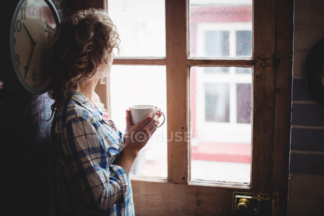 Жінка дивиться крізь вікно, маючи каву вдома — стокове фото