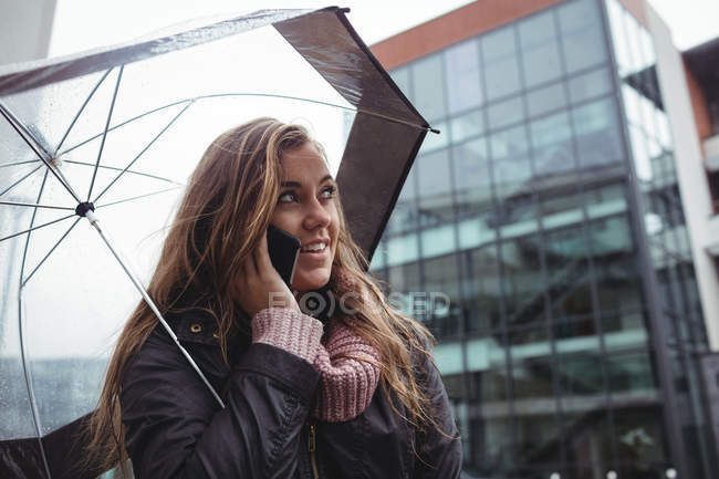 Belle femme tenant parapluie et parlant par smartphone sur la rue — Photo de stock