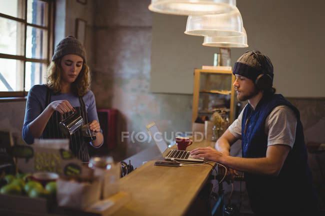 Serveuse préparant le café au comptoir tout en mécanicien utilisant un ordinateur portable en atelier — Photo de stock