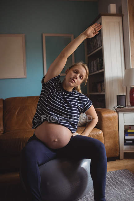 Mulher grávida realizando exercício de alongamento na bola de fitness na sala de estar em casa — Fotografia de Stock